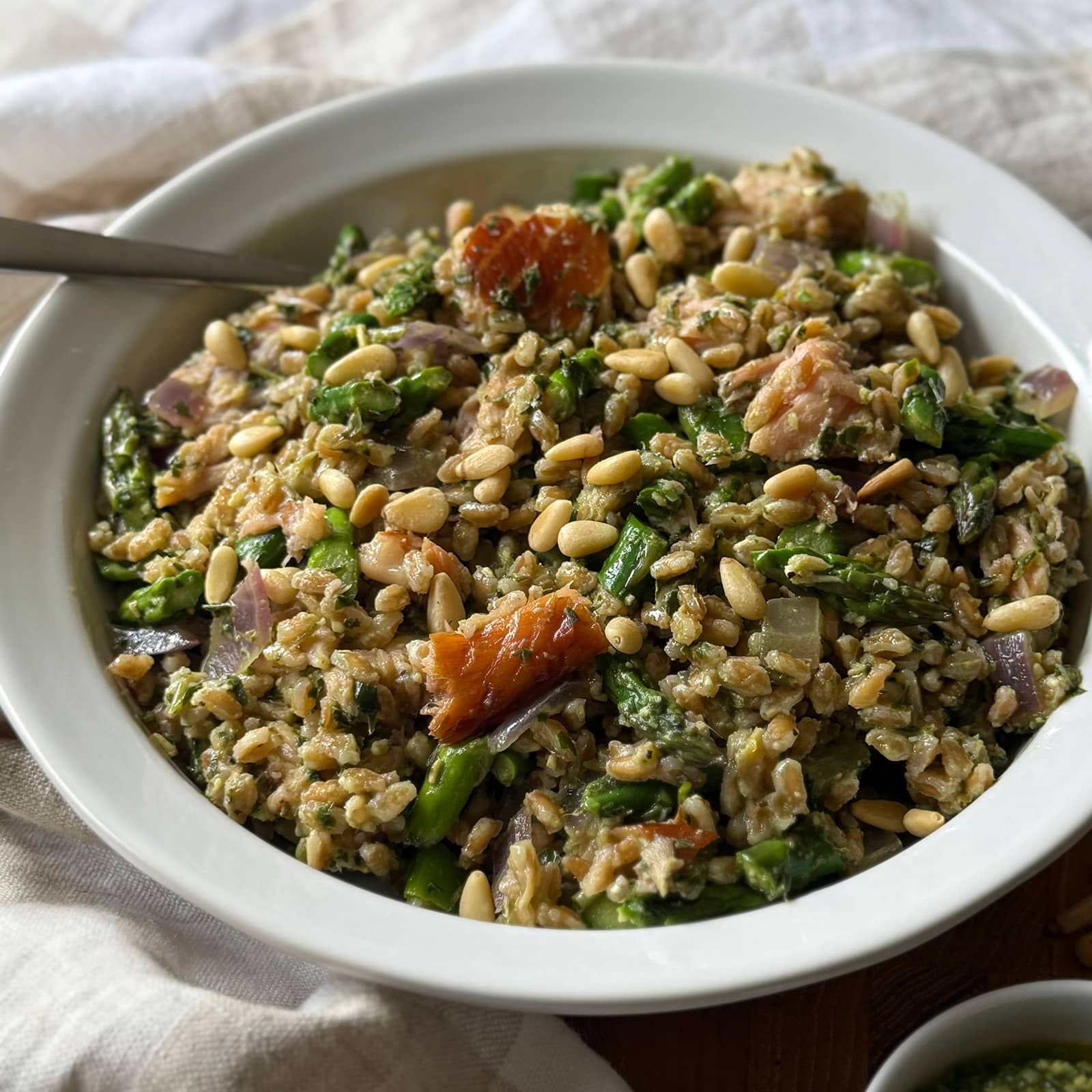 A bowl of farro, asparagus, red onions, pine nuts, and pieces of salmon, served with a fork. This Asparagus and Farro Salad with Salmon appears fresh and colorful, tossed with a fresh pesto sauce.
