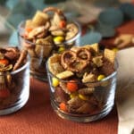 Three glass cups filled with Chex Mix for Fall, a blend of pretzels, Chex cereal, nuts, and colorful candy-coated chocolates are artfully arranged on an orange placemat.