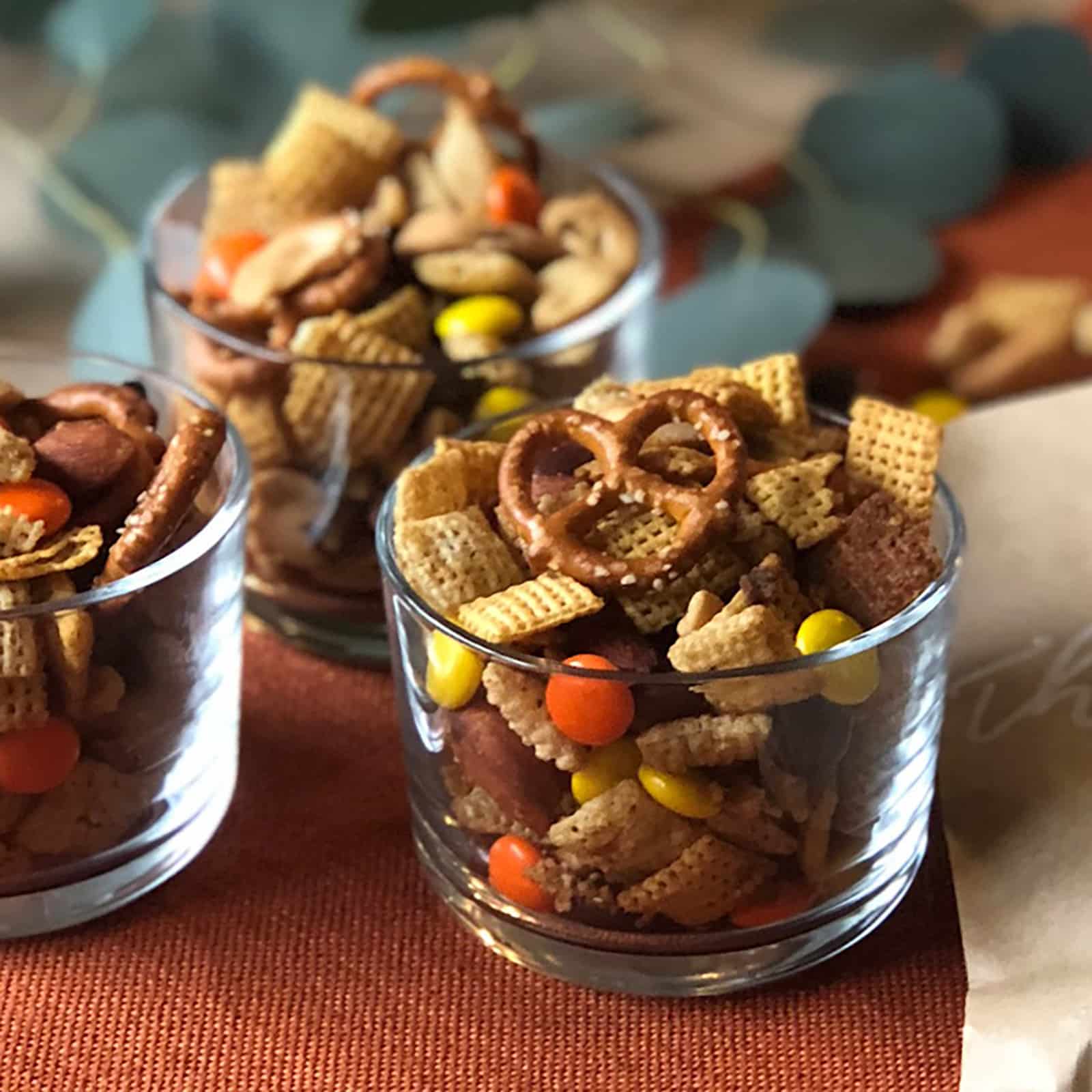 Three glass cups filled with Chex Mix for Fall, a blend of pretzels, Chex cereal, nuts, and colorful candy-coated chocolates are artfully arranged on an orange placemat.