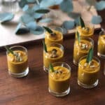 Small glass cups filled with Curried Pumpkin Soup, each topped with seeds and a sage leaf, are arranged on a wooden table with eucalyptus leaves in the background.