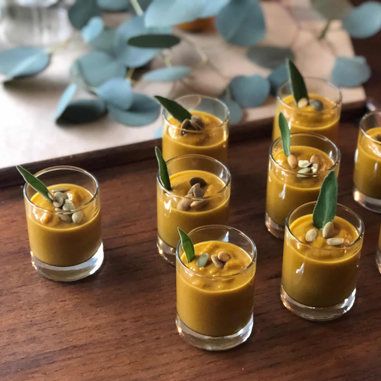 Small glass cups filled with Curried Pumpkin Soup, each topped with seeds and a sage leaf, are arranged on a wooden table with eucalyptus leaves in the background.