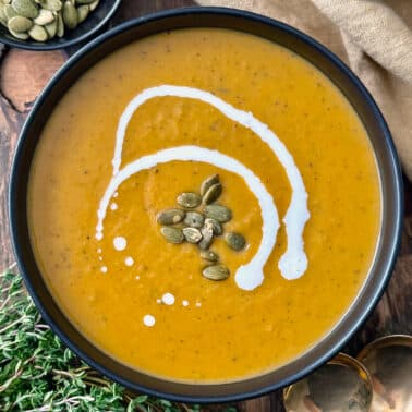 A bowl of creamy orange Acorn Squash Soup topped with pumpkin seeds and a swirl of coconut milk, set on a wooden table with herbs, a beige napkin, and a small dish of pumpkin seeds nearby.