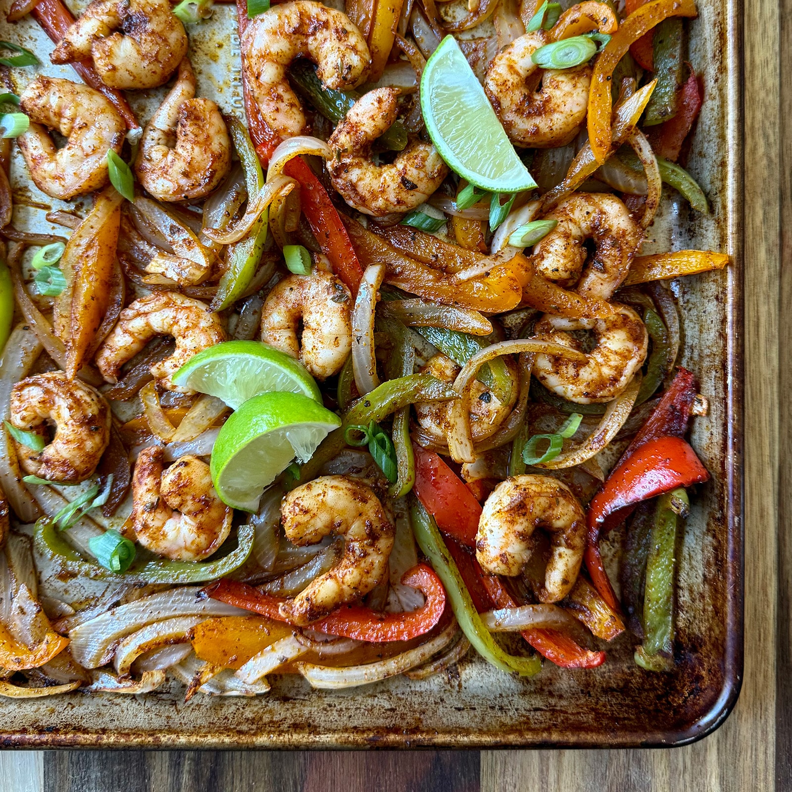 Sheet Pan Shrimp Fajitas featuring seasoned shrimp, sliced red and green bell peppers, onions, and lime wedges, all roasted together and garnished with chopped green onions on a wooden surface.