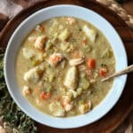 A bowl of creamy Seafood Chowder brimming with shrimp, chunks of white fish, carrots, celery, and fresh herbs, served in a white bowl with a spoon on a wooden surface.