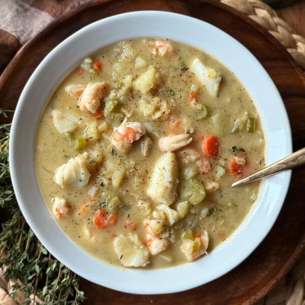A bowl of creamy Seafood Chowder brimming with shrimp, chunks of white fish, carrots, celery, and fresh herbs, served in a white bowl with a spoon on a wooden surface.