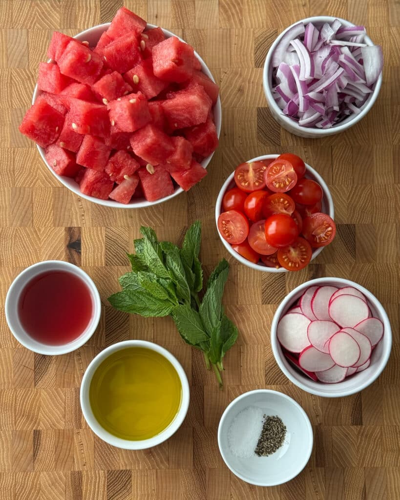 Ingredients for tomato and watermelon salad with radishes, including cubed watermelon, cherry tomatoes, sliced radishes, red onion, mint, olive oil, and red wine vinegar