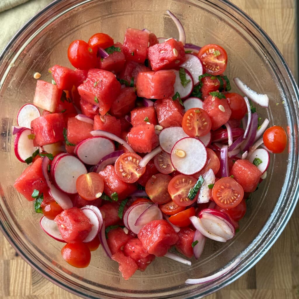 Tomato and watermelon salad with radishes being tossed together in a glass mixing bowl with fresh mint