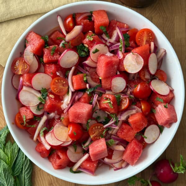 Overhead view of tomato and watermelon salad with radishes in a white bowl, garnished with fresh mint and flaked salt