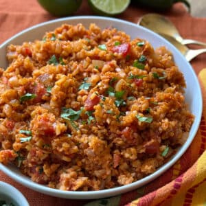 A bowl of Spanish Cauliflower Rice garnished with chopped cilantro, featuring visible diced tomatoes, onions, and spices, is placed on an orange cloth with two gold spoons and limes in the background.