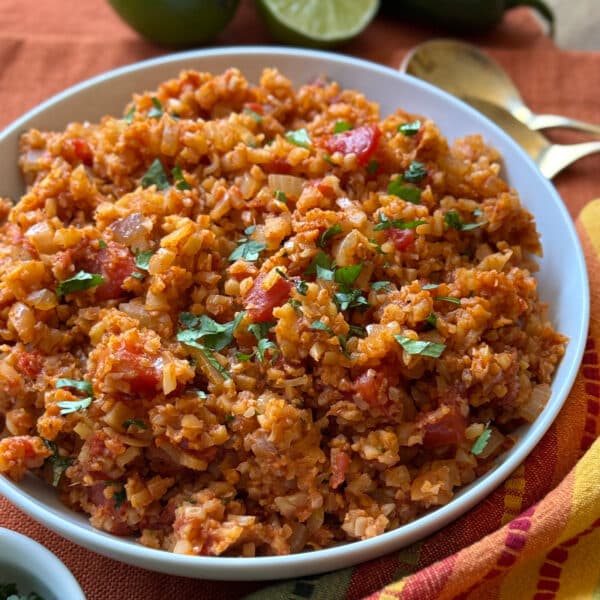 A bowl of Spanish Cauliflower Rice garnished with chopped cilantro, featuring visible diced tomatoes, onions, and spices, is placed on an orange cloth with two gold spoons and limes in the background.