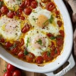 Roasted Tomatoes with Baked Eggs features perfectly baked eggs with runny yolks nestled in a dish of roasted cherry tomatoes, garnished with chopped basil and a sprinkle of black pepper.