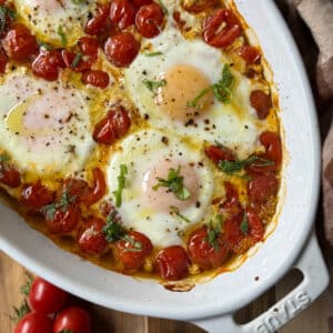 Roasted Tomatoes with Baked Eggs features perfectly baked eggs with runny yolks nestled in a dish of roasted cherry tomatoes, garnished with chopped basil and a sprinkle of black pepper.