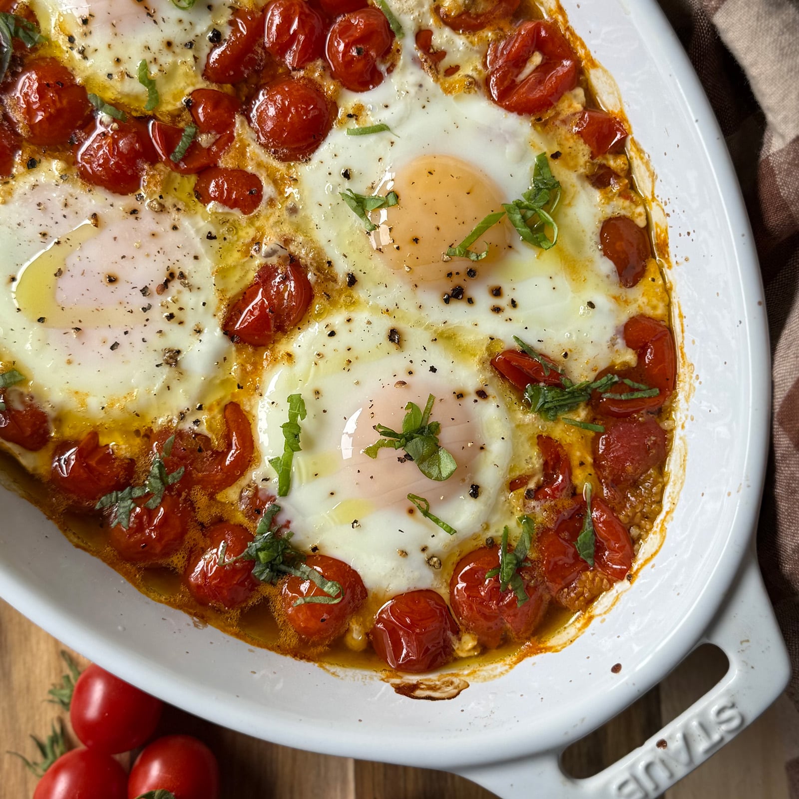 Roasted Tomatoes with Baked Eggs features perfectly baked eggs with runny yolks nestled in a dish of roasted cherry tomatoes, garnished with chopped basil and a sprinkle of black pepper.