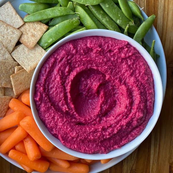 A plate with vibrant beet hummus, crackers, sugar snap peas, and baby carrots sits on a wooden table adorned with fresh lemons, blue napkins, white plates, and a jar of Ruby Red Kraut.