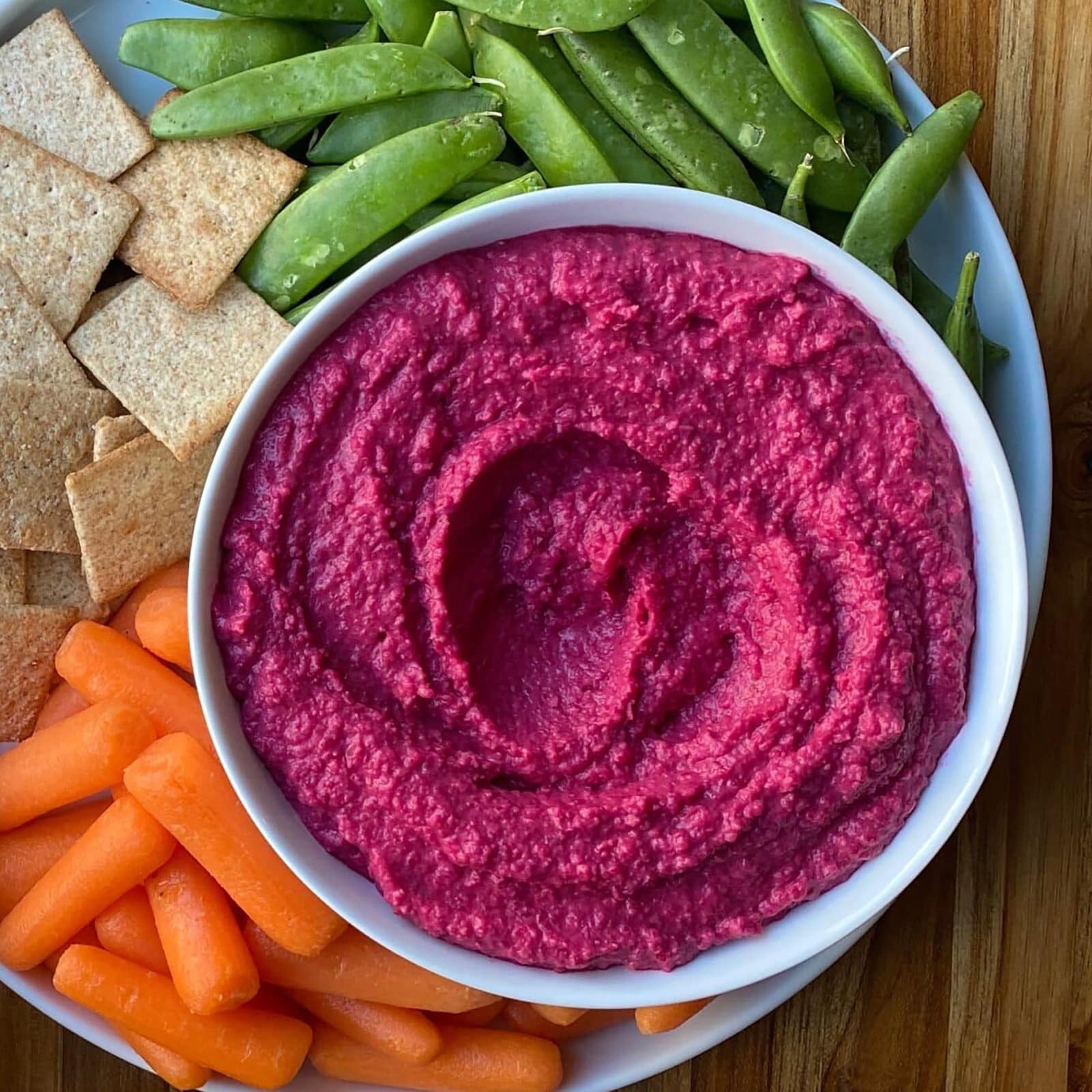 A plate with vibrant beet hummus, crackers, sugar snap peas, and baby carrots sits on a wooden table adorned with fresh lemons, blue napkins, white plates, and a jar of Ruby Red Kraut.