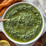 A white bowl filled with vibrant green carrot top pesto sauce, with a spoon inside. Roasted carrots and a lemon wedge are partially visible next to the bowl on a white plate.