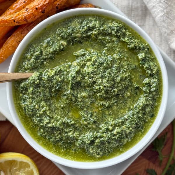 Top-down view of carrot top pesto in a white bowl with a wooden spoon, surrounded by roasted carrots and a halved lemon