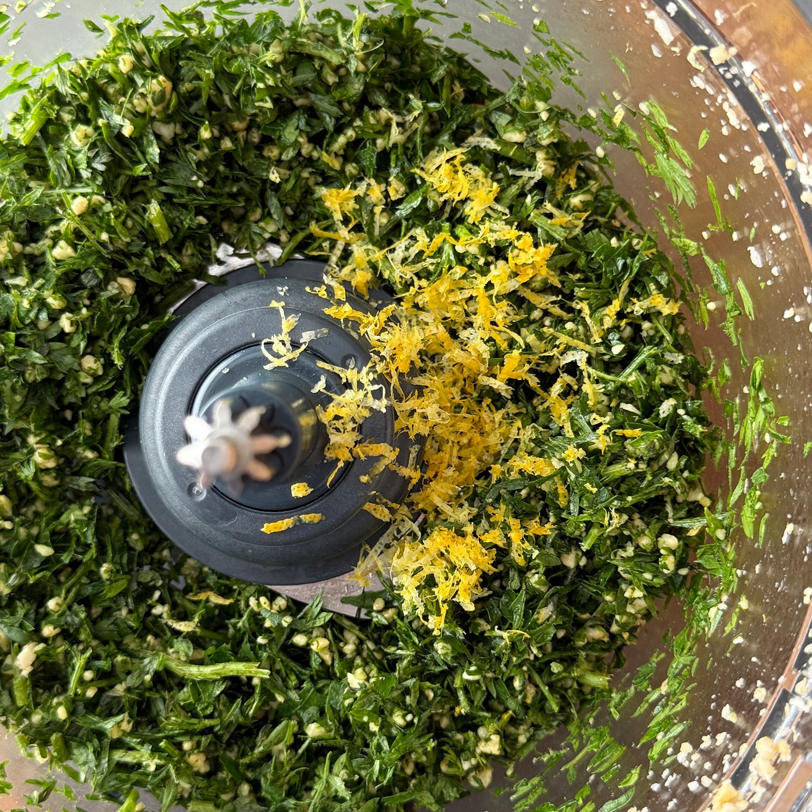 Chopped carrot tops and parsley topped with fresh lemon zest in a food processor