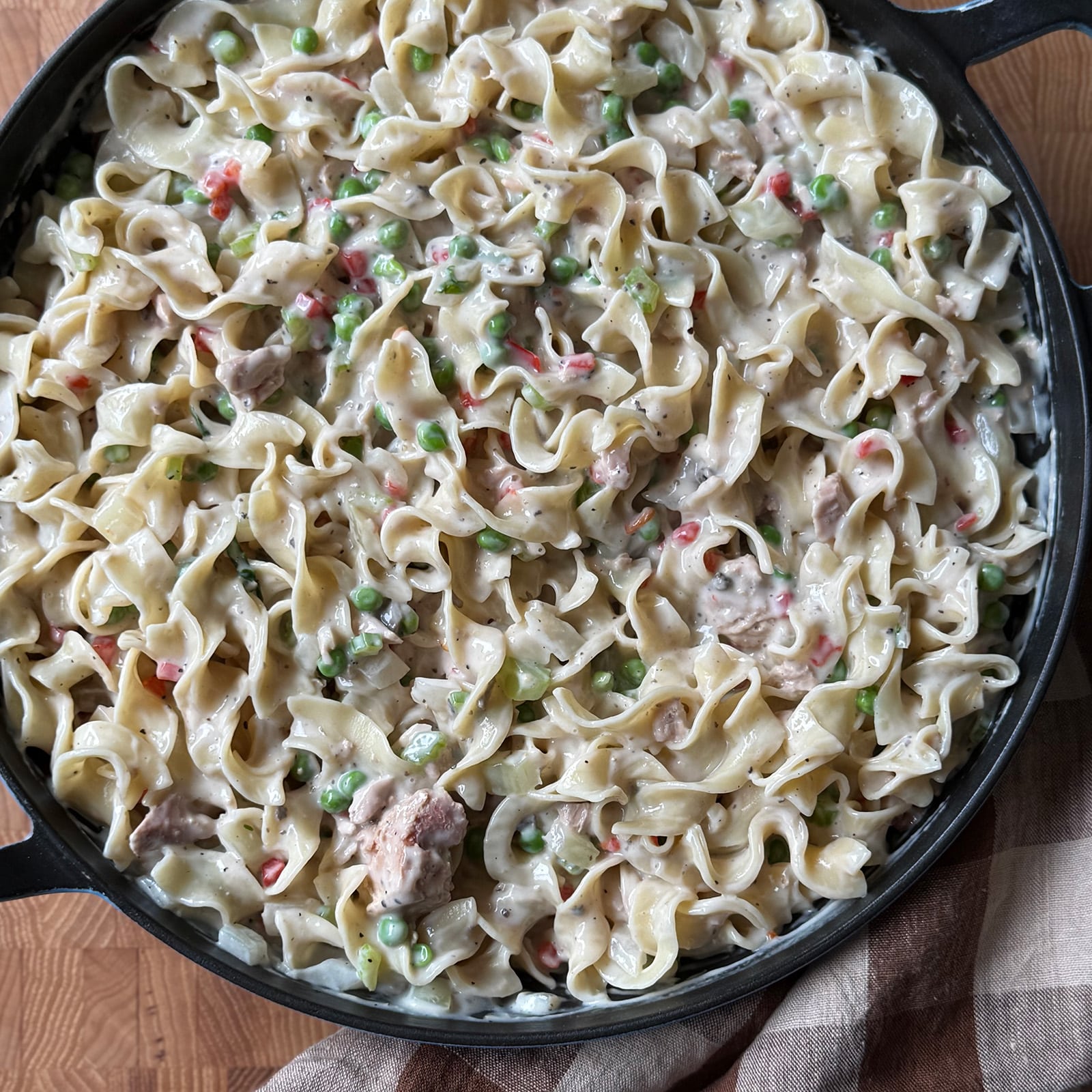 Tuna noodle casserole filling with egg noodles, peas, tuna, and diced pimentos in a cast iron skillet