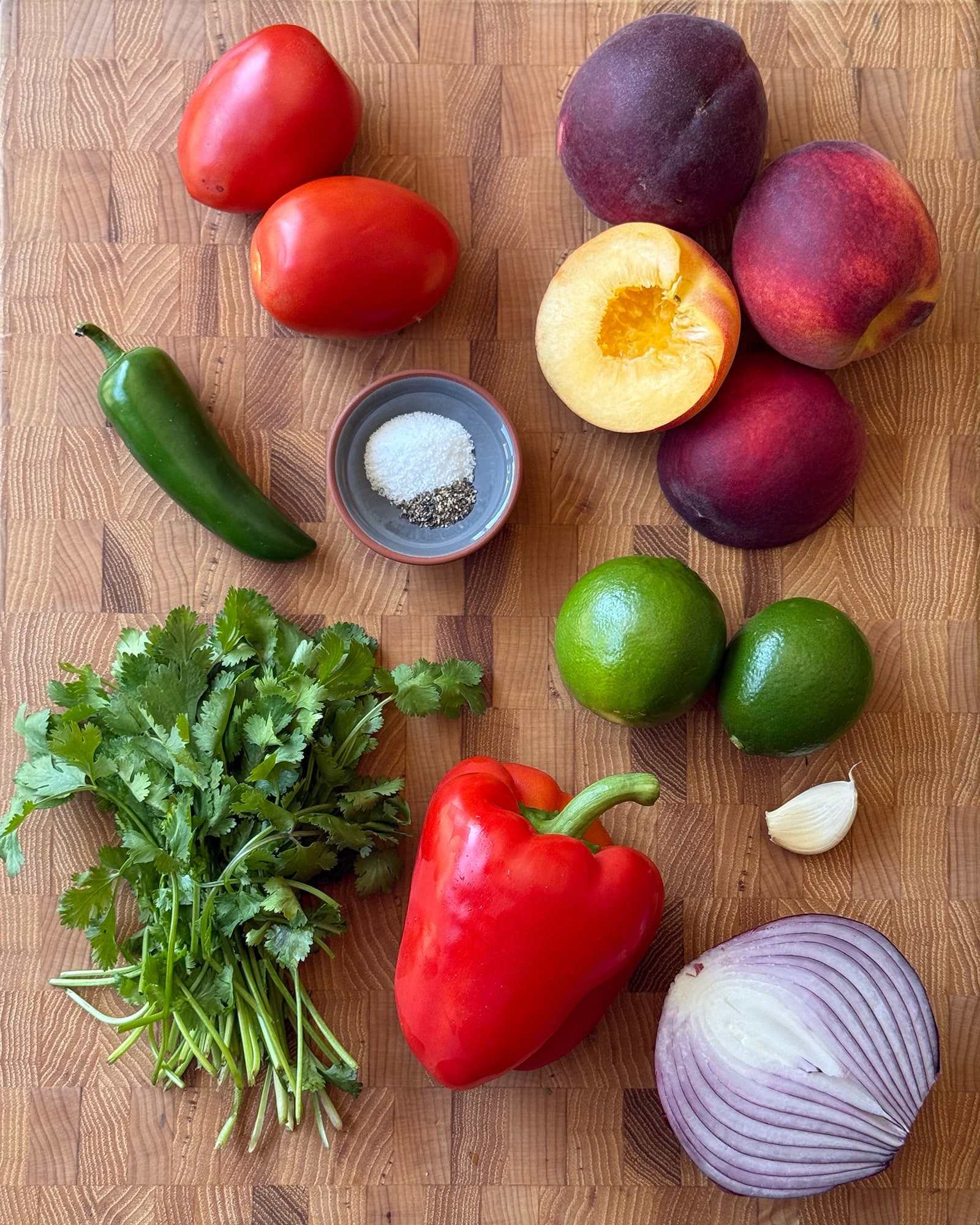 Flat lay of peach salsa ingredients on a butcher block cutting board including whole and halved peaches, roma tomatoes, a red bell pepper, jalape&ntilde;o, halved red onion, fresh cilantro, two limes, a garlic clove, and a small dish of salt and pepper