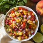 Overhead view of peach salsa dip in a white bowl on a wooden surface surrounded by fresh cilantro, a whole peach, and halved limes, showing diced peaches, red bell pepper, tomato, red onion, and cilantro
