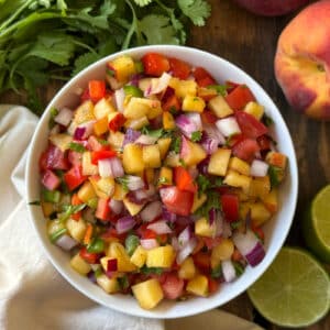 Overhead view of peach salsa dip in a white bowl on a wooden surface surrounded by fresh cilantro, a whole peach, and halved limes, showing diced peaches, red bell pepper, tomato, red onion, and cilantro