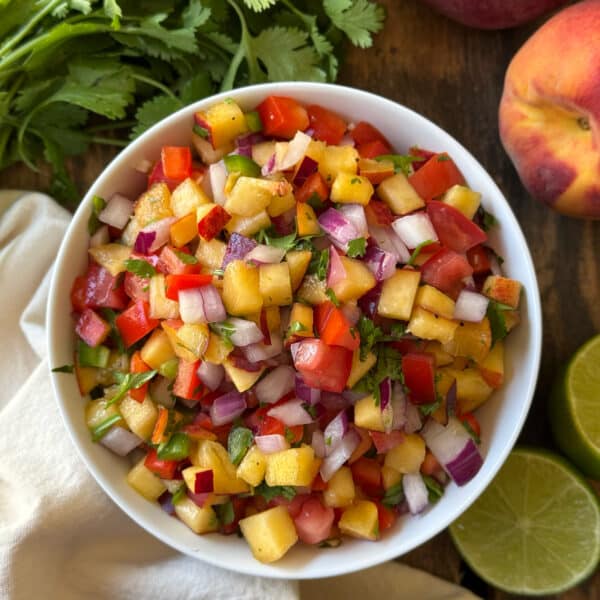 Overhead view of peach salsa dip in a white bowl on a wooden surface surrounded by fresh cilantro, a whole peach, and halved limes, showing diced peaches, red bell pepper, tomato, red onion, and cilantro