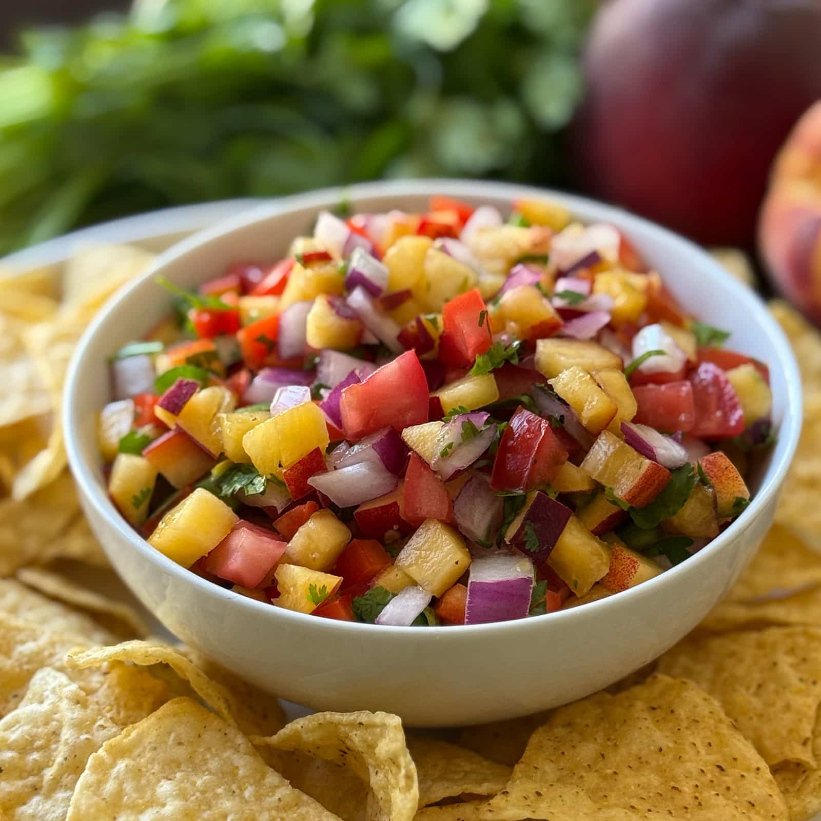 Angled shot of peach salsa dip in a white bowl surrounded by tortilla chips with fresh cilantro and a whole peach visible in the background