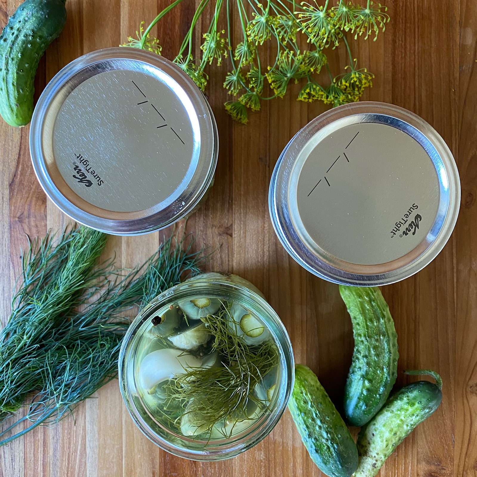 Overhead view of three mason jars of dill pickles on a wooden cutting board &mdash; two sealed with silver lids and one open showing fresh dill, garlic, and peppercorns &mdash; surrounded by fresh dill flowers and whole pickling cucumbers