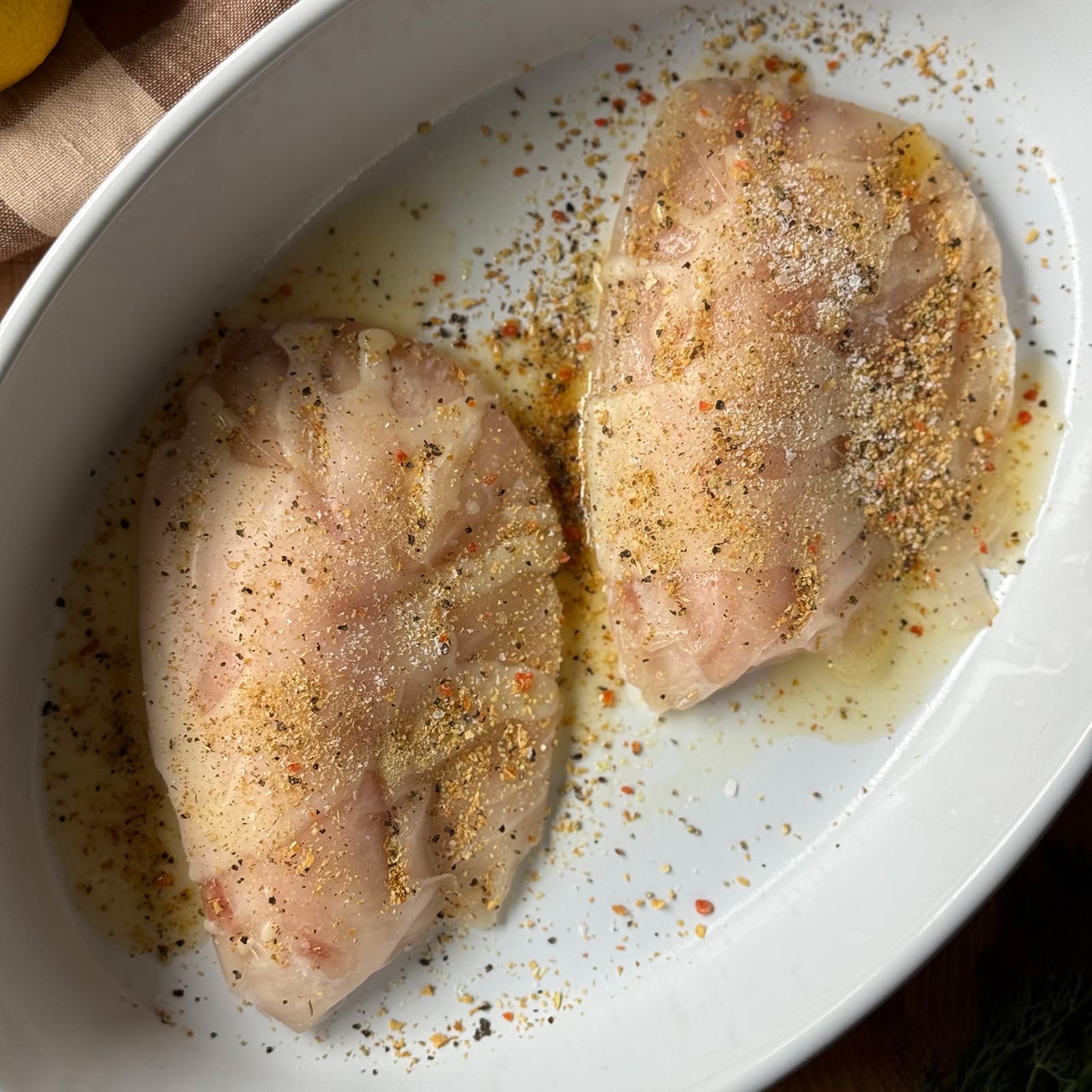 Two frozen tilapia fillets in a white oval baking dish seasoned with lemon pepper blend and kosher salt, ready to bake