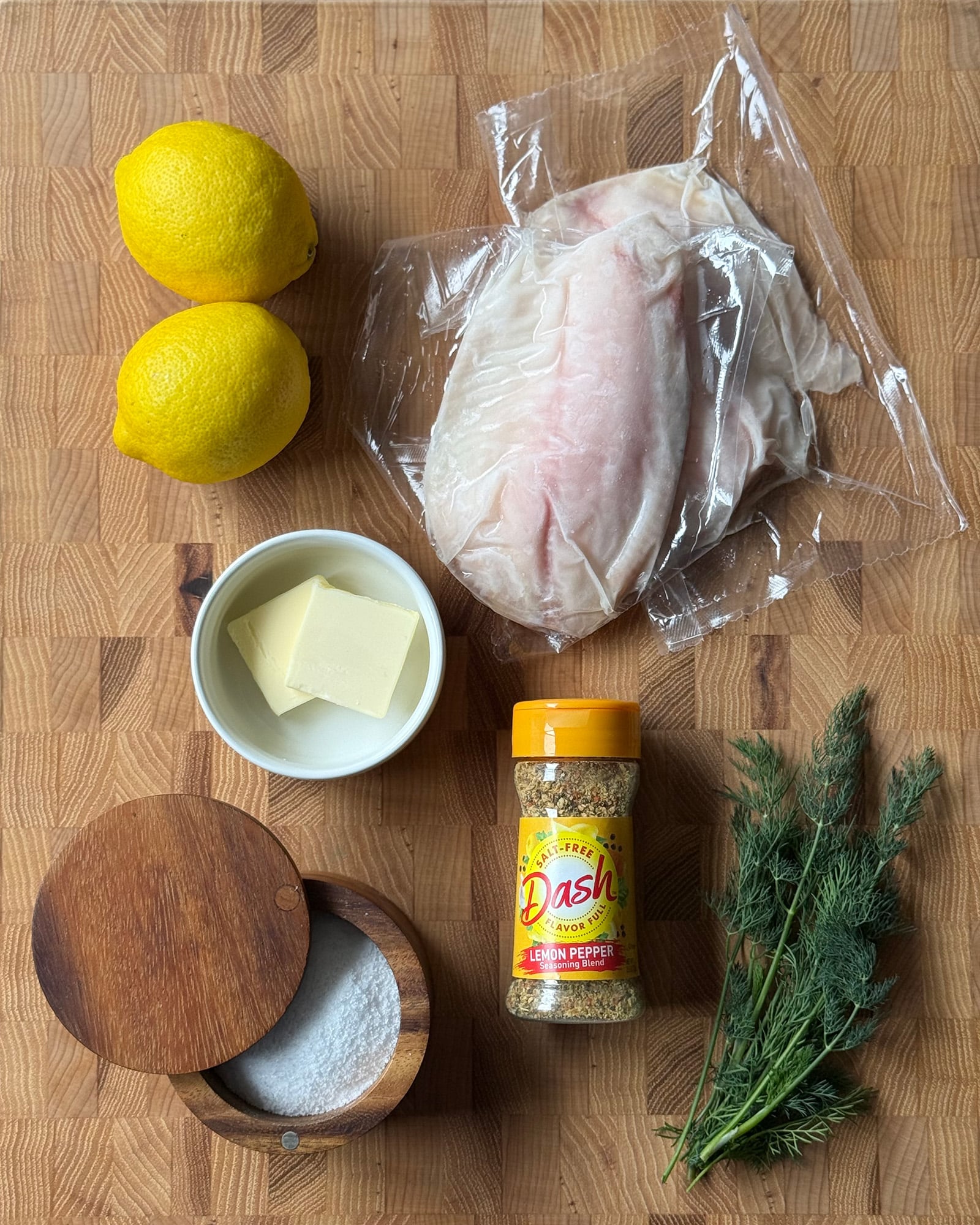 Ingredients for lemon pepper tilapia on a wood cutting board including frozen tilapia fillets, lemons, butter, Dash lemon pepper seasoning, kosher salt, and fresh dill