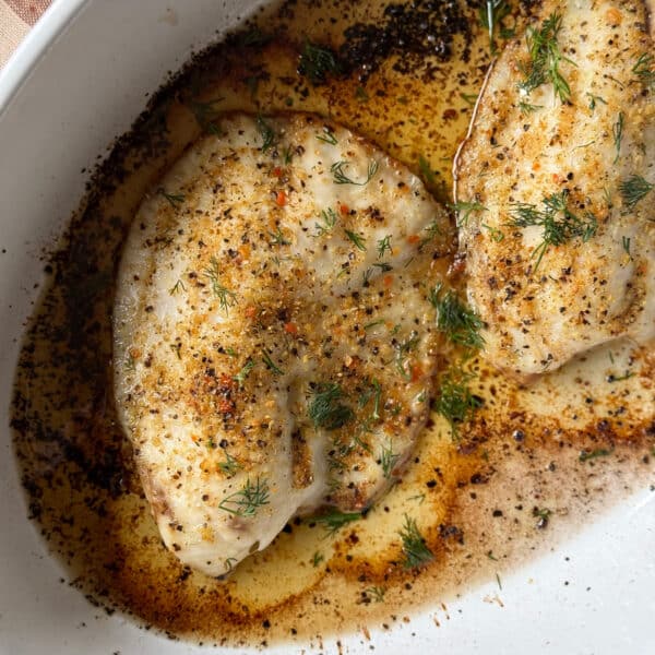 Two lemon pepper tilapia fillets baked in a white oval dish, topped with fresh dill and surrounded by golden butter sauceTwo lemon pepper tilapia fillets baked in a white oval dish, topped with fresh dill and surrounded by golden butter sauce