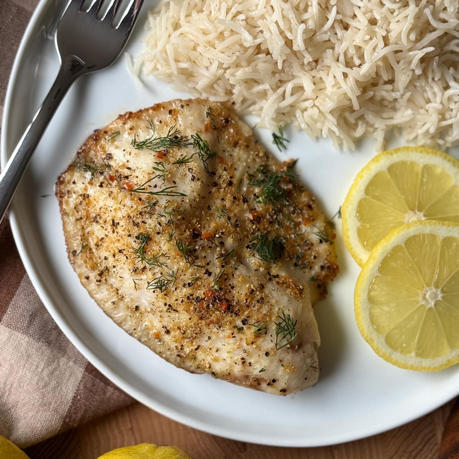 A plated lemon pepper tilapia fillet served with basmati rice and lemon slices on a white plate with fresh dill and lemon slices