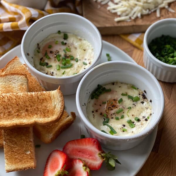 Two white ramekins of baked eggs with melted Gruyere, runny yolks, cracked black pepper, and fresh chives on a plate with toast strips and halved strawberries, with shredded Gruyere on a wooden board behind.