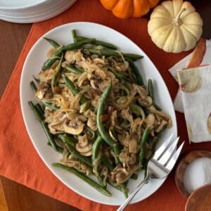 An oval platter of Green Beans with Mushroom & Onion sits on a table draped with an orange cloth. The cooked green beans are elegantly paired with saut&eacute;ed mushrooms and onions, creating a vibrant, autumn-inspired side dish. Small pumpkins, plates, and a folded napkin add seasonal charm nearby, while a serving fork rests invitingly on the platter.