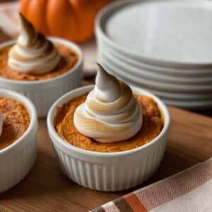 A small sweet potato casserole in a white ramekin, topped with swirled toasted marshmallow fluff. Stacked plates and a pumpkin are in the background on a wooden surface.