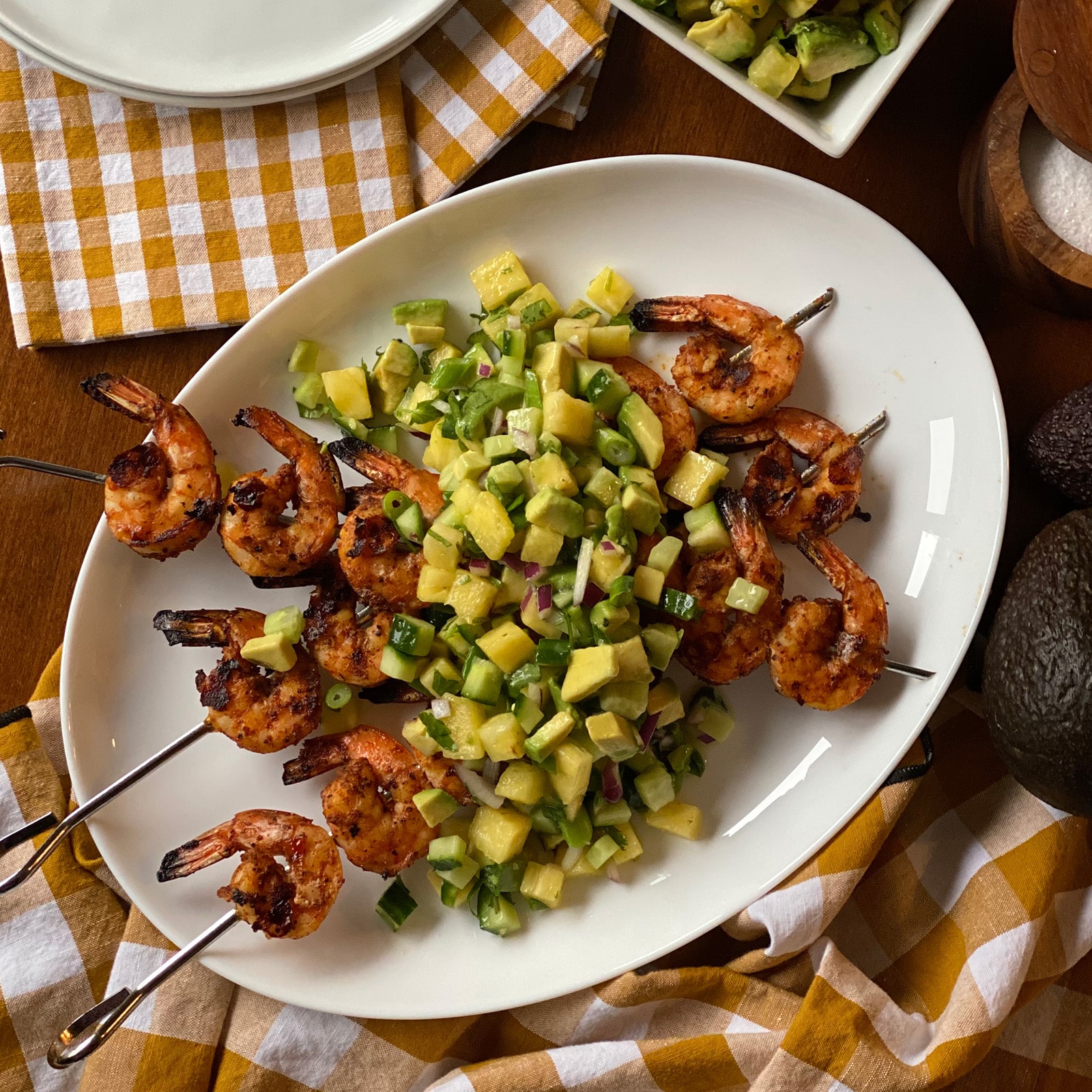 Overhead view of grilled shrimp skewers arranged on a white oval platter with pineapple cucumber avocado salsa in the center, surrounded by a gold checkered linen and fresh avocados