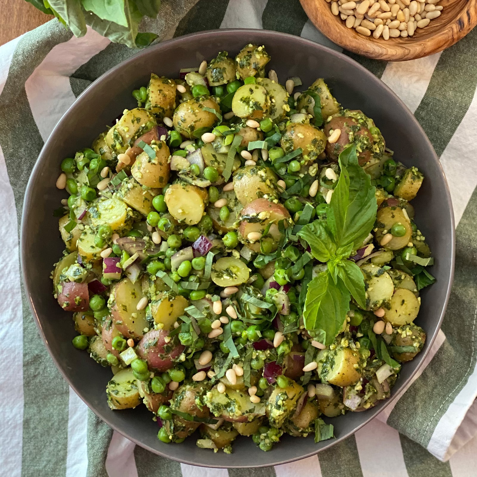 Overhead view of pesto potato salad in a gray bowl with fingerling potatoes, green peas, red onion, toasted pine nuts, and fresh basil, on a striped linen napkin.
