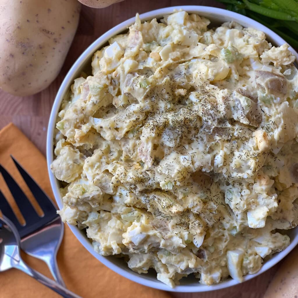 Overhead view of classic potato salad with hard-boiled eggs, celery, and dried dill in a white bowl on a wooden cutting board.