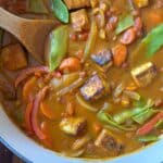 A wooden spoon stirs a curry filled with cubes of seared tofu, sliced carrots, snow peas, and red bell pepper strips in a thick, orange-brown sauce.
