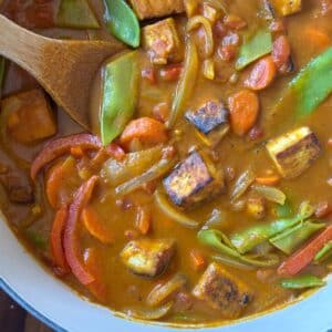 A wooden spoon stirs a curry filled with cubes of seared tofu, sliced carrots, snow peas, and red bell pepper strips in a thick, orange-brown sauce.