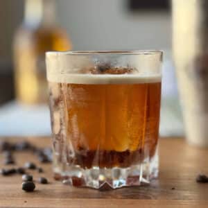 A carajillo cocktail served over a large ice cube in a chilled rocks glass, garnished with three whole coffee beans