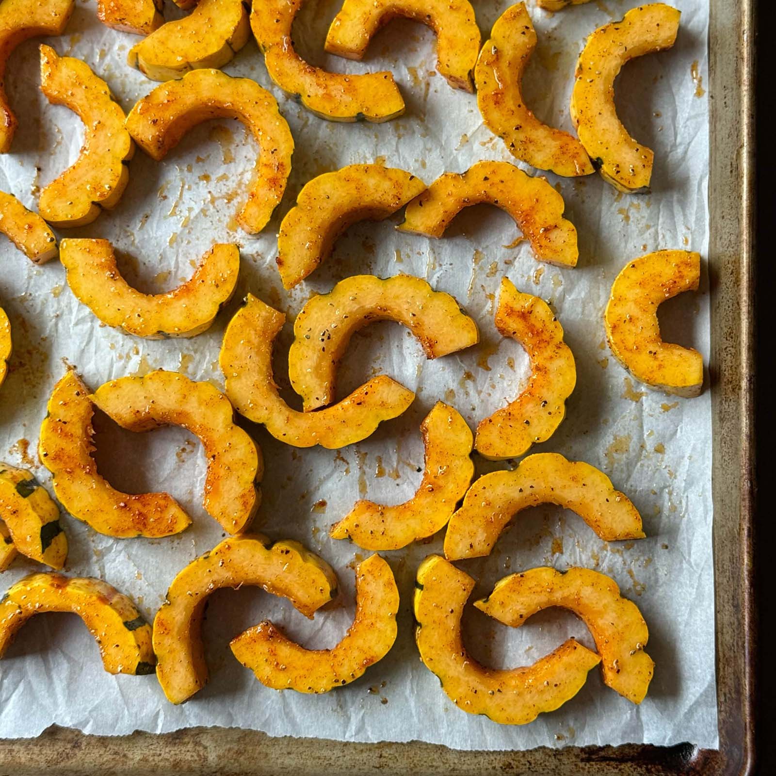 Baked Delicata Squash pieces are sliced and arranged on a parchment-lined baking sheet, then roasted until seasoned and golden brown.