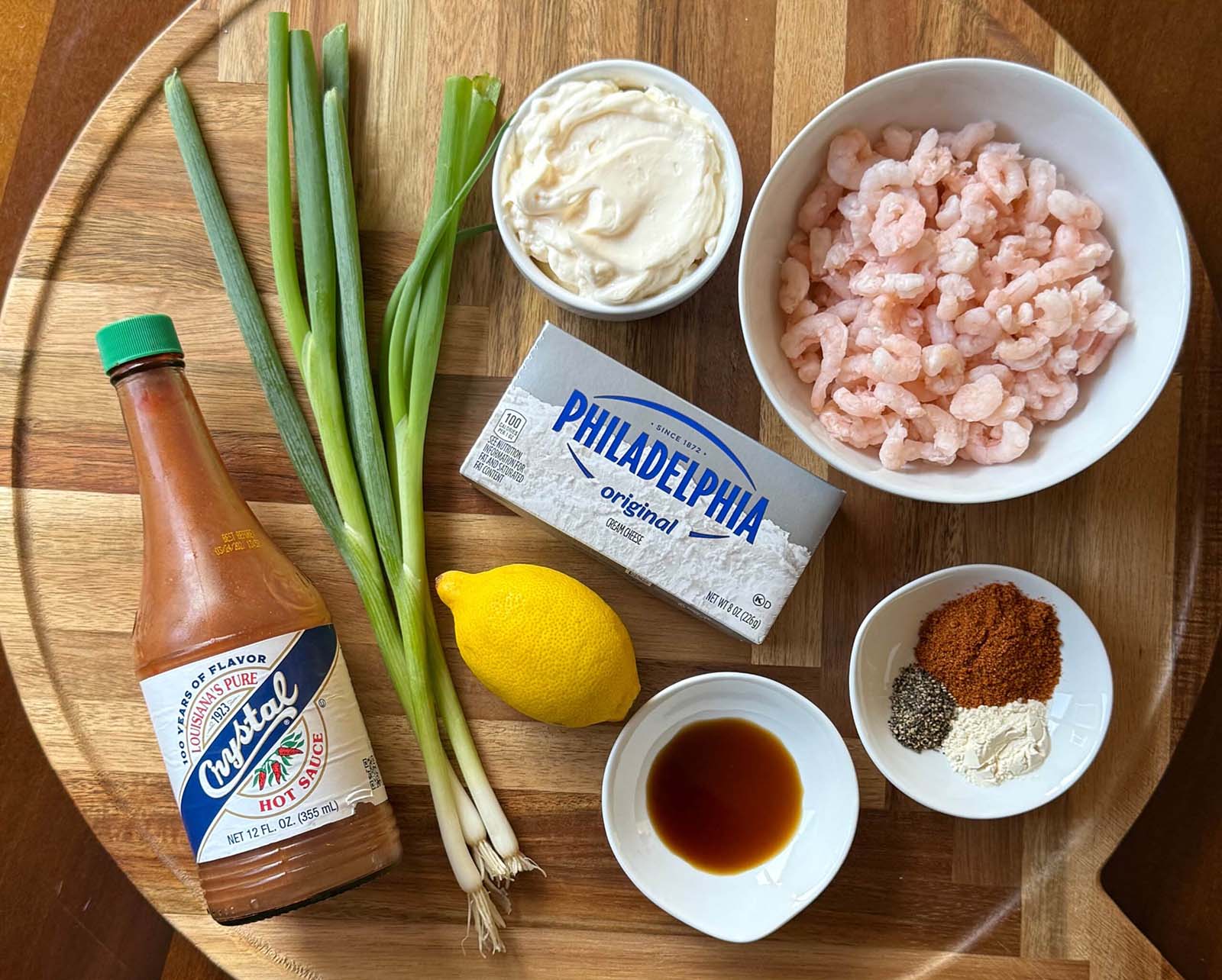 Ingredients for creamy shrimp dip arranged on a wooden tray, including cream cheese, mayonnaise, frozen salad shrimp, Old Bay seasoning, lemon, green onions, and hot sauce.
