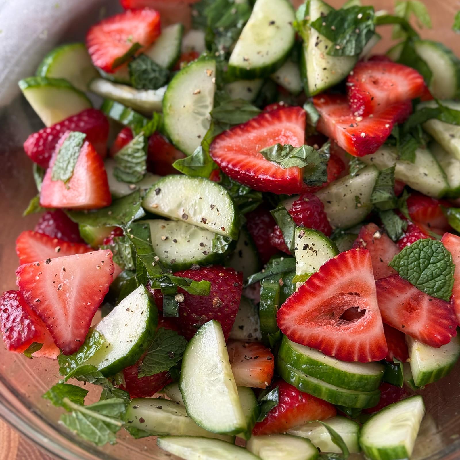 Close-up of cucumber and strawberry salad showing sliced strawberries, cucumber, and fresh mint and basil leaves glistening with lime dressing and cracked black pepper
