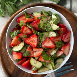 Overhead view of cucumber and strawberry salad in a white bowl on a wooden serving board with a striped linen tablecloth and serving fork.