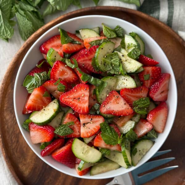 Overhead view of cucumber and strawberry salad in a white bowl on a wooden serving board with a striped linen tablecloth and serving fork.