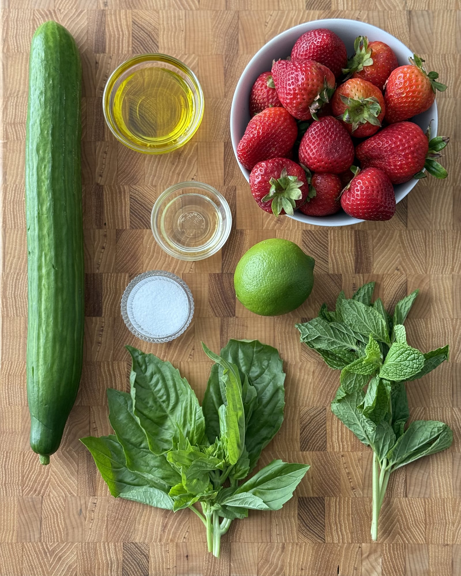 Flat lay of cucumber and strawberry salad ingredients on a butcher block cutting board including a whole English cucumber, fresh strawberries in a white bowl, olive oil, agave, a lime, kosher salt, fresh basil, and fresh mint