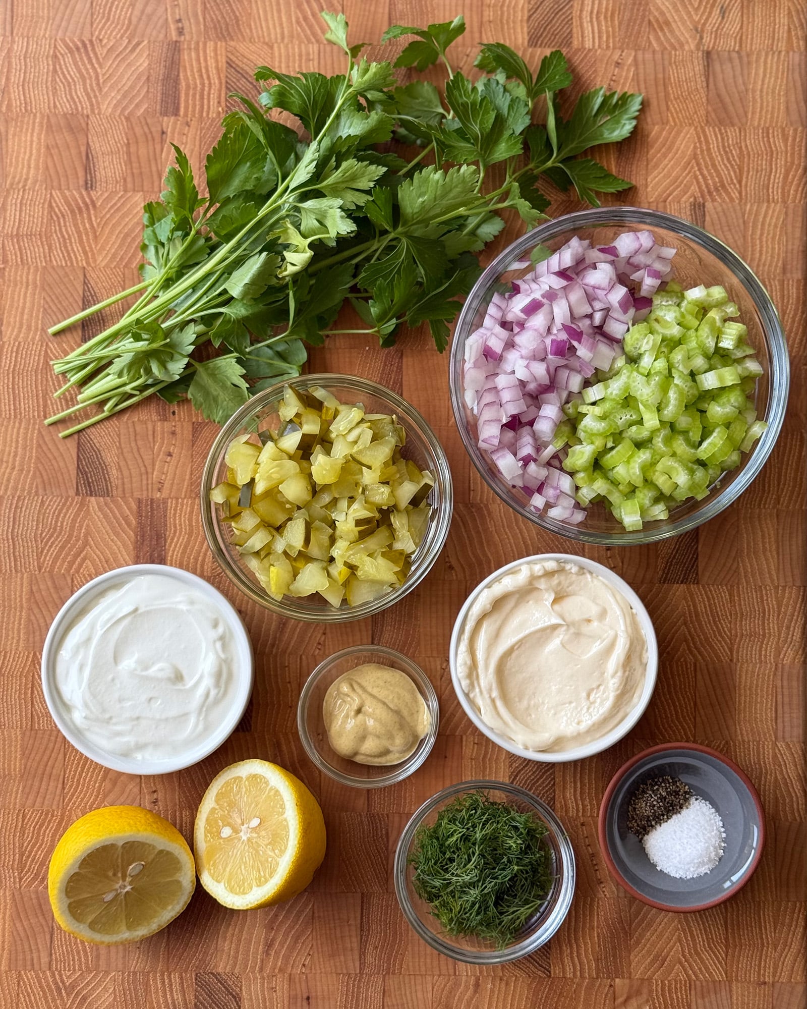 Ingredients for crispy tater tot potato salad including fresh parsley, dill, chopped dill pickles, red onion, celery, mayonnaise, Greek yogurt, Dijon mustard, lemon, and kosher salt arranged on a butcher block cutting board.