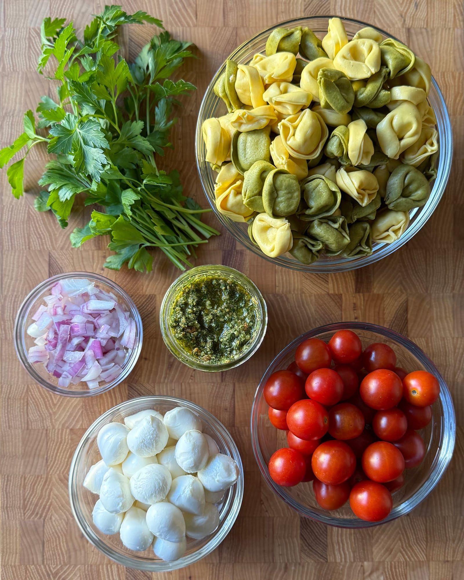 Ingredients for Tortellini Pasta Salad with Pesto arranged in glass bowls on a butcher block surface, including tortellini, cherry tomatoes, mini mozzarella, red onion, fresh parsley, and a jar of mint pesto]